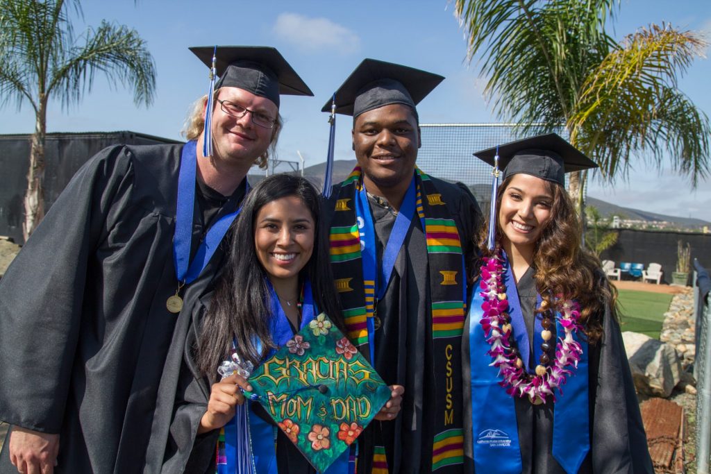 4 graduates in cap in gown from CSUSM looking at the camera 